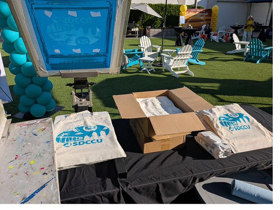 Live screen printing setup with printed SDCCU tote bags at a corporate event in San Diego